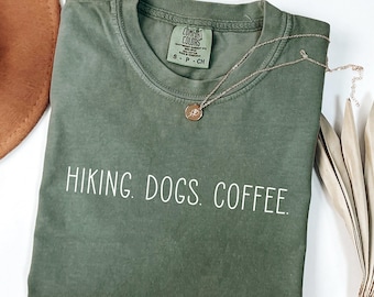 May include: An olive green t-shirt with the words "HIKING. DOGS. COFFEE." in white. A gold necklace with a pendant rests on the shirt. The shirt has a Comfort Colors tag.