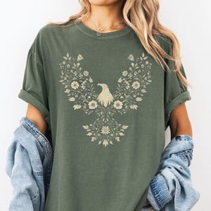 May include: Olive green t-shirt featuring a floral eagle design. The eagle is composed of flowers and leaves in a cream color. The shirt has short sleeves and a relaxed fit, perfect for casual wear.