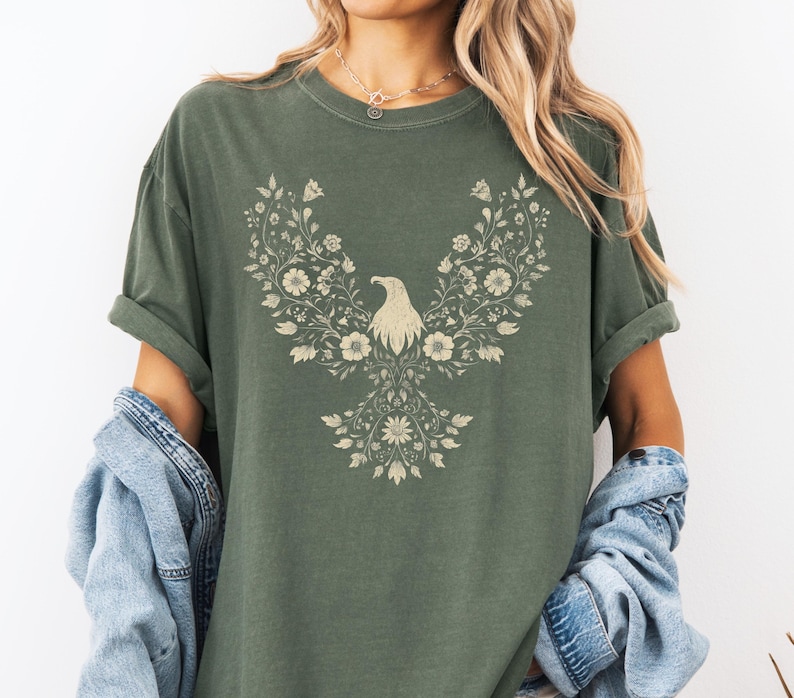 May include: Olive green t-shirt featuring a floral eagle design. The eagle is composed of flowers and leaves in a cream color. The shirt has short sleeves and a relaxed fit, perfect for casual wear.