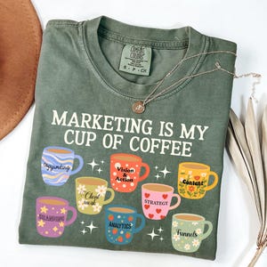 May include: An olive green short-sleeve t-shirt featuring the white text "MARKETING IS MY CUP OF COFFEE" above eight colorful illustrated mugs. Each mug displays a specific marketing term including Copywriting, Vision & Action, Content, Client work, Strategy, Branding, Analytics, and Funnels.