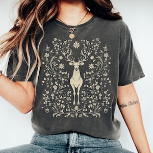 May include: Dark gray t-shirt featuring a cream-colored deer design surrounded by floral elements. The deer has large antlers and stands centered on the shirt. The shirt has a crew neck and short sleeves.