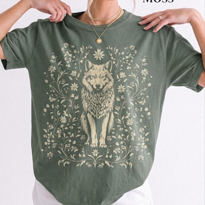 May include: A moss green short-sleeve t-shirt featuring a cream-colored graphic print of a wolf centered among intricate floral vines and leaves. The text MOSS is printed in the upper right corner of the image above the crew neck collar.