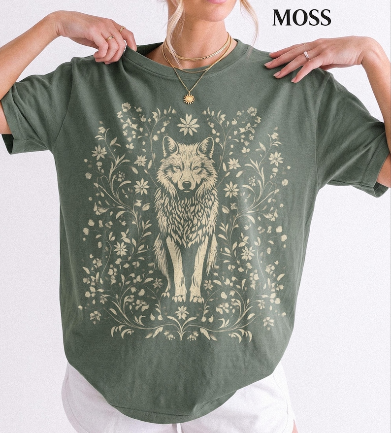 May include: A moss green short-sleeve t-shirt featuring a cream-colored graphic print of a wolf centered among intricate floral vines and leaves. The text MOSS is printed in the upper right corner of the image above the crew neck collar.