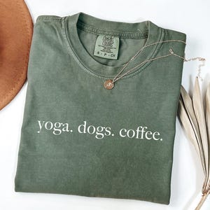 May include: Olive green t-shirt with the words "yoga. dogs. coffee." in white. A gold necklace with a pendant rests on the shirt. The shirt is folded and has a tag that says "Comfort Colors".