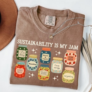 May include: A brown Comfort Colors t-shirt with the text "SUSTAINABILITY IS MY JAM" and illustrations of jars labeled "Eco living", "Recycling", "Reuse & repair", "Clean water", "Green energy", "Plant based", and "Composting". A silver necklace is on the shirt.