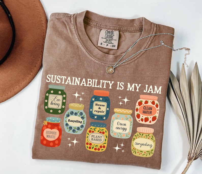 May include: A brown Comfort Colors t-shirt with the text "SUSTAINABILITY IS MY JAM" and illustrations of jars labeled "Eco living", "Recycling", "Reuse & repair", "Clean water", "Green energy", "Plant based", and "Composting". A silver necklace is on the shirt.