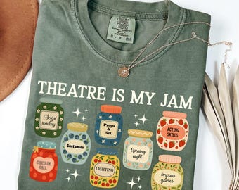 May include: Sage green t-shirt with the phrase "THEATRE IS MY JAM" and illustrations of jam jars labeled with theater-related terms like "Script reading" and "Acting Skills." A silver necklace with a pendant is draped across the shirt.