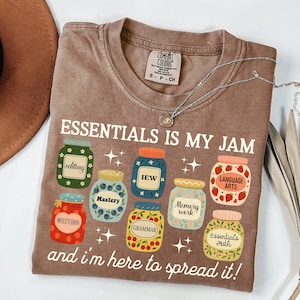 May include: A brown Comfort Colors t-shirt with the text "ESSENTIALS IS MY JAM" and various jar graphics, each labeled with educational terms like "editing" and "language arts." The shirt also includes the phrase "and I'm here to spread it!"