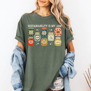 May include: Green t-shirt with the text "SUSTAINABILITY IS MY JAM" above illustrated jars of food. The jars are in various colors and designs, with labels like "I'm berry", "Clean Water", and "Plant Based".