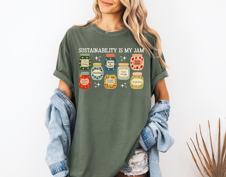 May include: Green t-shirt with the text "SUSTAINABILITY IS MY JAM" above illustrated jars of food. The jars are in various colors and designs, with labels like "I'm berry", "Clean Water", and "Plant Based".