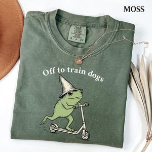 May include: A moss green crew neck t-shirt featuring a graphic of a frog wearing a pointed hat and riding a kick scooter. The text "Off to train dogs" is printed in white above the frog. The shirt has a Comfort Colors brand label at the neck.