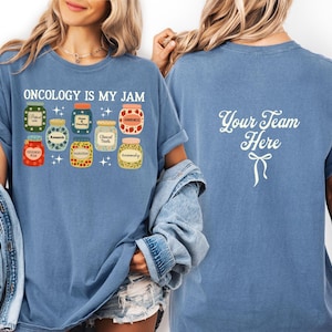 May include: A blue t-shirt with the text "ONCOLOGY IS MY JAM" and a graphic of various jam jars. The back of the shirt has the text "Your Team Here" with a bow graphic. The shirt is made of a soft material.