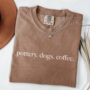 May include: A brown t-shirt with the words "pottery. dogs. coffee." printed in white. A silver necklace with a small pendant is draped across the shirt. A brown hat and dried plant are in the background.