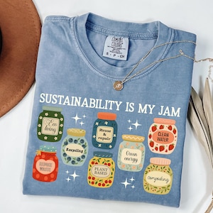 May include: Light blue Comfort Colors t-shirt with the text "SUSTAINABILITY IS MY JAM" and illustrations of jars with eco-friendly labels. The jars have labels such as "Eco living", "Recycling", "Clean Water", and "Composting". A gold necklace is draped across the shirt.