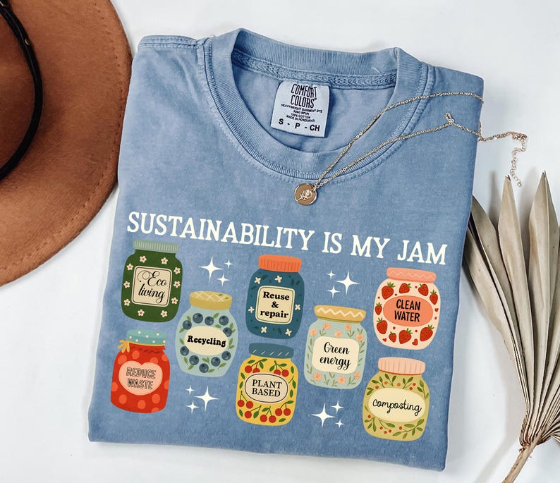 May include: Light blue Comfort Colors t-shirt with the text "SUSTAINABILITY IS MY JAM" and illustrations of jars with eco-friendly labels. The jars have labels such as "Eco living", "Recycling", "Clean Water", and "Composting". A gold necklace is draped across the shirt.
