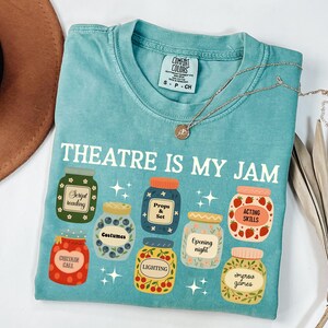 May include: A turquoise Comfort Colors t-shirt with the text "THEATRE IS MY JAM" and illustrations of jam jars labeled with theater-related terms like "Script Reading" and "Acting Skills." A gold necklace is draped across the shirt.