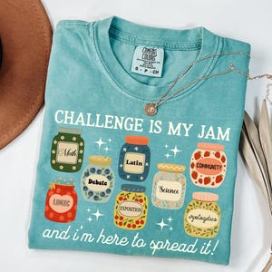 May include: A turquoise Comfort Colors t-shirt with the text "CHALLENGE IS MY JAM" and the phrase "and I'm here to spread it!" The shirt features jar graphics with words like "Math", "Latin", and "Community". A gold necklace is draped on the shirt.