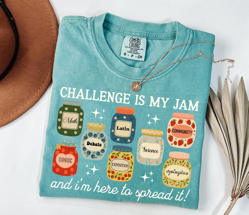 May include: A turquoise Comfort Colors t-shirt with the text "CHALLENGE IS MY JAM" and the phrase "and I'm here to spread it!" The shirt features jar graphics with words like "Math", "Latin", and "Community". A gold necklace is draped on the shirt.