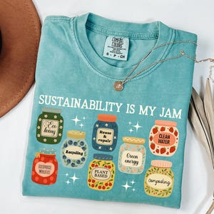 May include: A turquoise t-shirt with the text "SUSTAINABILITY IS MY JAM" and illustrations of jars with eco-friendly labels. The shirt has a crew neck and short sleeves. A gold necklace with a pendant is on the shirt.