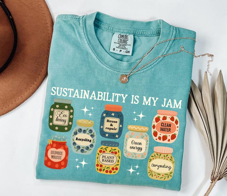 May include: A turquoise t-shirt with the text "SUSTAINABILITY IS MY JAM" and illustrations of jars with eco-friendly labels. The shirt has a crew neck and short sleeves. A gold necklace with a pendant is on the shirt.