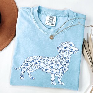 May include: Light blue Comfort Colors t-shirt featuring a floral dachshund design in white and blue. The shirt has a crew neck and short sleeves. A silver necklace with a small pendant is draped across the shirt.