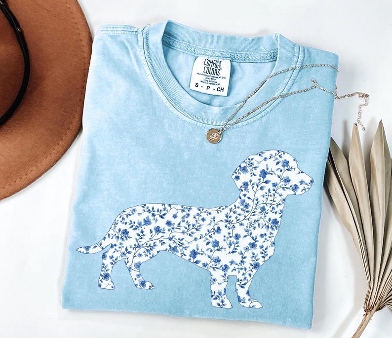 May include: Light blue Comfort Colors t-shirt featuring a floral dachshund design in white and blue. The shirt has a crew neck and short sleeves. A silver necklace with a small pendant is draped across the shirt.