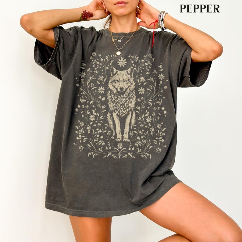 May include: A charcoal gray short-sleeve t-shirt featuring a central graphic of a wolf surrounded by intricate floral and vine illustrations. The design is printed in a light, muted tone, contrasting with the dark fabric of the shirt.