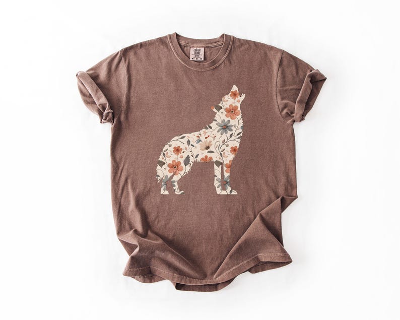 May include: A brown t-shirt featuring a floral wolf design. The wolf silhouette is filled with a pattern of flowers in shades of orange, cream, and green. The shirt has a crew neck and short sleeves, with a slightly faded appearance.
