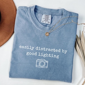 May include: A blue Comfort Colors t-shirt with the text "easily distracted by good lighting" and a camera graphic. A gold necklace with a pendant rests on the shirt. A brown hat and dried plant are in the background.