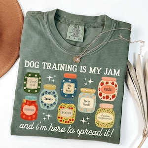 May include: Green Comfort Colors t-shirt with the phrase "DOG TRAINING IS MY JAM" and illustrations of jars labeled with dog training commands. A silver necklace and a brown hat are also visible.