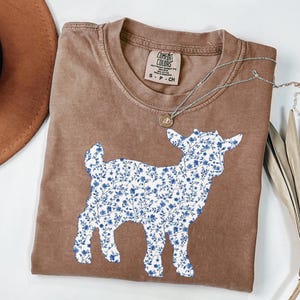 May include: Brown t-shirt with a goat silhouette filled with a blue floral pattern. The shirt has a crew neck and a small tag near the collar. A silver necklace with a small pendant is draped across the shirt.