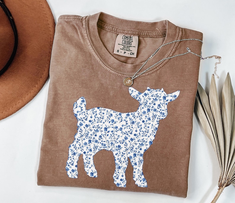 May include: Brown t-shirt with a goat silhouette filled with a blue floral pattern. The shirt has a crew neck and a small tag near the collar. A silver necklace with a small pendant is draped across the shirt.