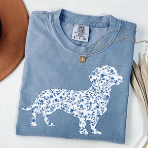 May include: A blue Comfort Colors t-shirt featuring a floral dachshund design. The dog silhouette is filled with a blue and white floral pattern. A gold necklace with a pendant rests on the shirt. The shirt is size S-P-CH.
