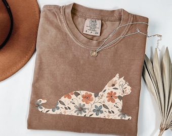 May include: A brown Comfort Colors t-shirt featuring a floral cat design in shades of orange, blue, and cream. The shirt has a crew neck and short sleeves. A silver necklace with a small pendant is draped across the shirt.