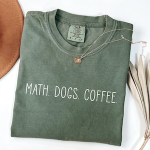 May include: Olive green t-shirt with the words "MATH. DOGS. COFFEE." in white. A gold necklace with a pendant rests on the shirt. The shirt is folded and placed on a white surface.