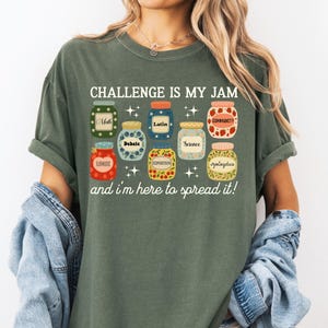 May include: Olive green t-shirt with the text "CHALLENGE IS MY JAM" and the phrase "and I'm here to spread it!" below. The shirt features colorful jar illustrations with labels like "Math", "Latin", and "Community".