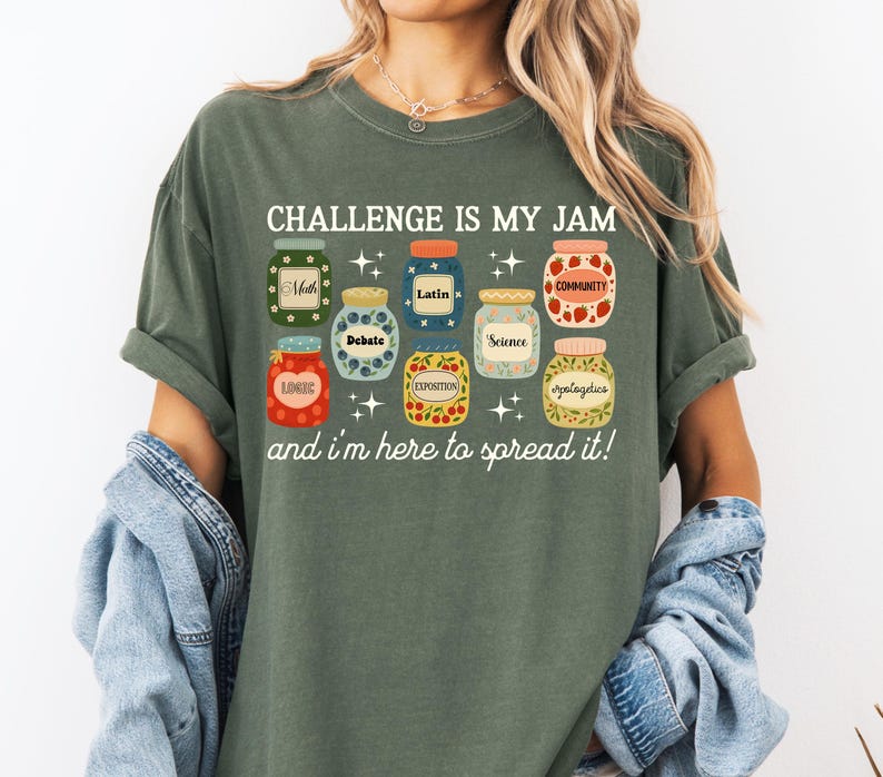 May include: Olive green t-shirt with the text "CHALLENGE IS MY JAM" and the phrase "and I'm here to spread it!" below. The shirt features colorful jar illustrations with labels like "Math", "Latin", and "Community".