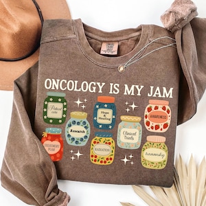 May include: A brown long-sleeved sweatshirt with the text "ONCOLOGY IS MY JAM" in white. The design includes illustrated jars with labels like "Patient Care," "Research," and "Awareness." A necklace and a hat are also visible.