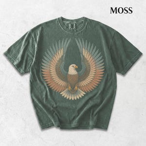 May include: A moss green short-sleeve t-shirt featuring a graphic of a bald eagle with wings spread wide. The eagle is rendered in shades of brown, tan, and muted blue. The word MOSS is printed in the upper right corner of the image.