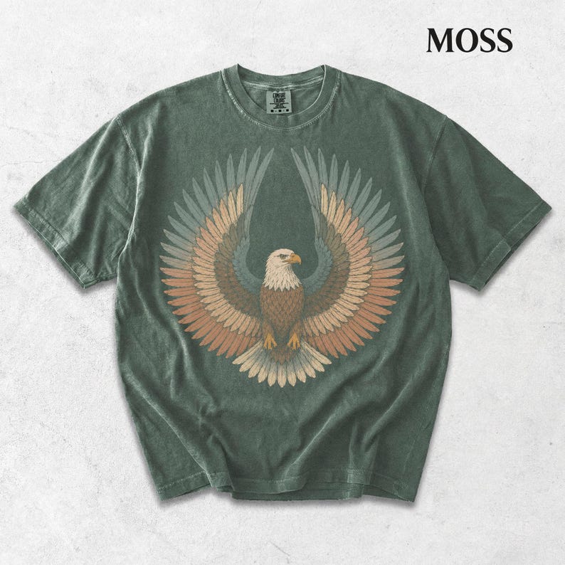 May include: A moss green short-sleeve t-shirt featuring a graphic of a bald eagle with wings spread wide. The eagle is rendered in shades of brown, tan, and muted blue. The word MOSS is printed in the upper right corner of the image.