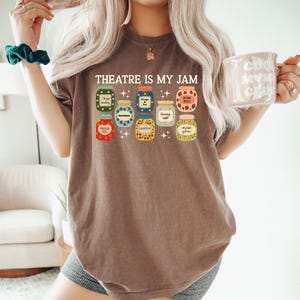 May include: Brown t-shirt with the text "THEATRE IS MY JAM" above a graphic of various jam jars. Each jar has a different label, including "Bright Reading" and "Acting Skills".