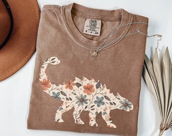 Könnte beinhalten: Ein braunes Comfort Colors T-Shirt mit einem floralen Dinosaurier-Design. Der Dinosaurier ist mit orangefarbenen, blauen und cremefarbenen Blumen gefüllt. Eine silberne Halskette mit einem kleinen Anhänger liegt auf dem Shirt.