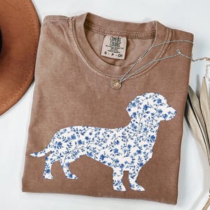 May include: A brown t-shirt featuring a floral dachshund design in blue and white. The shirt has a crew neck and short sleeves. A silver necklace with a small pendant is on the shirt.