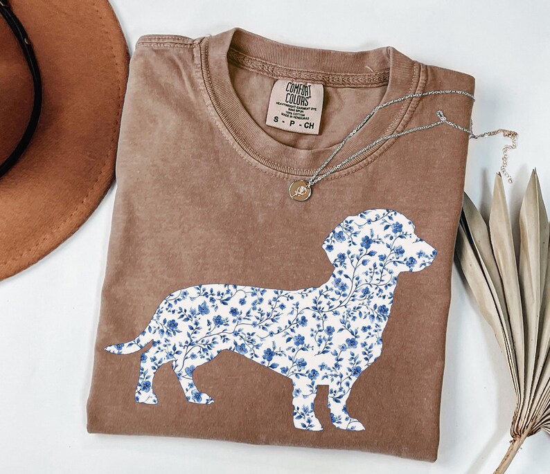 May include: A brown t-shirt featuring a floral dachshund design in blue and white. The shirt has a crew neck and short sleeves. A silver necklace with a small pendant is on the shirt.