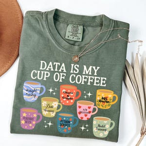 May include: An olive green t-shirt featuring the white text DATA IS MY CUP OF COFFEE above seven colorful mugs. Each mug displays a data-related label including Insight Mining, Charts & Graphs, SQL Queries, Data Cleaning, A/B Testing, Feature Sets, Python Scripts, and Model Training.