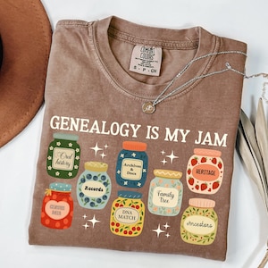 May include: A brown t-shirt with the phrase "GENEALOGY IS MY JAM" and illustrations of jam jars. The jars have labels like "Oral History," "Records," and "Heritage." A silver necklace rests on the shirt.