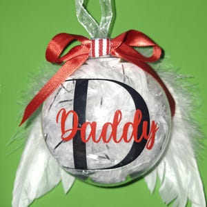 Könnte beinhalten: Eine transparente Weihnachtskugel, gefüllt mit weißen Federn und silbernem Lametta. Die Kugel hat ein rotes Band und das Wort "Daddy" in roter Schrift. Ein großer schwarzer Buchstabe "D" befindet sich hinter dem Wort "Daddy". Weiße Federn sind an den Seiten befestigt.