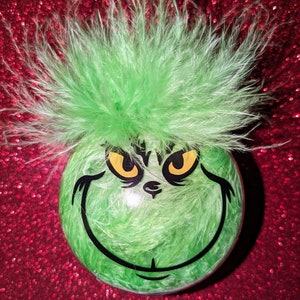 Könnte beinhalten: Grüner Weihnachtsbaumschmuck mit einem flauschigen grünen Grinch-Gesicht und gelben Augen. Der Schmuck ist mit schwarzem Besatz verziert und liegt auf einem roten, glitzernden Hintergrund.