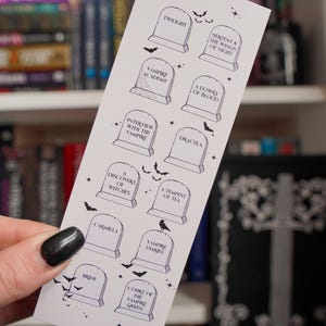 May include: A white bookmark with tombstone illustrations and book titles such as "Twilight" and "Dracula" printed in black. Small bat and star illustrations are scattered around the tombstones. The bookmark is held in a hand.