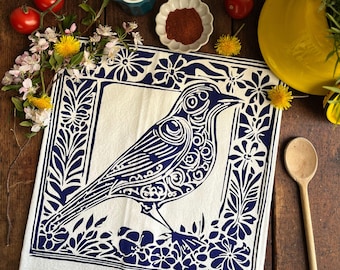 Puede incluir: Una toalla de cocina blanca con un borde floral azul y un pájaro estilizado azul en el centro. El pájaro está detallado con intrincados patrones y está posado en un lecho de flores.